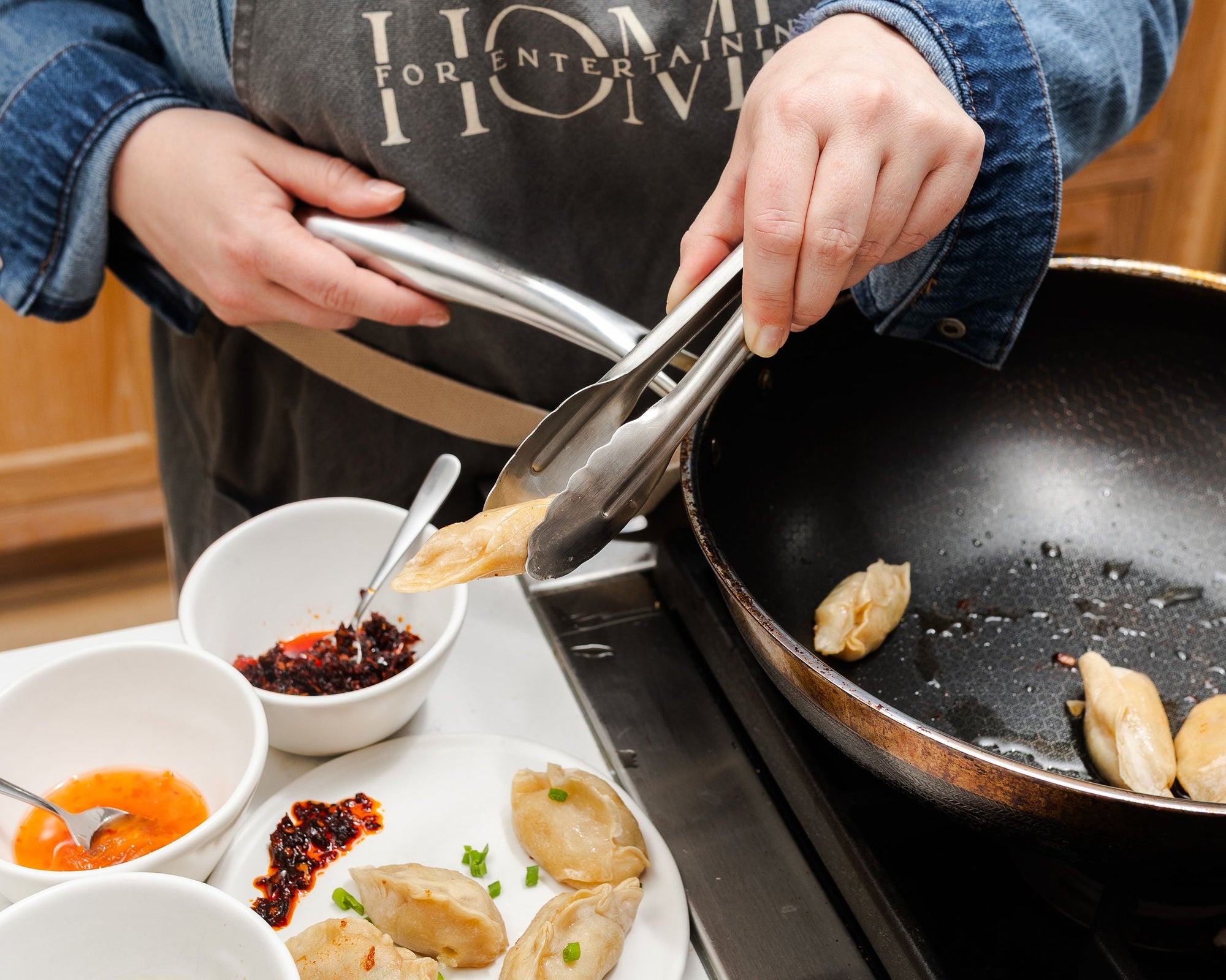 Person cooking potstickers in a frying pan with various sauces on a counter.